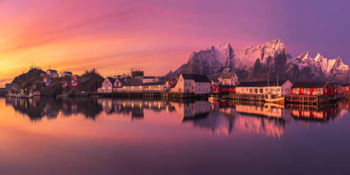 Atemberaubender Blick auf das Dorf Reine mit seinen roten und weißen Häusern an der Küste des ruhigen Meeres vor dem Hintergrund des verschneiten Bergrückens und des Sonnenuntergangs auf den Lofoten, Norwegen - ADSF33705