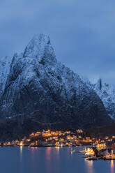 Spektakulärer Blick auf hohe schneebedeckte Berge und ein Dorf mit beleuchteten Häusern in einer Bucht am Fuße eines Hügels in Reine auf den Lofoten in Norwegen bei Sonnenuntergang - ADSF33706