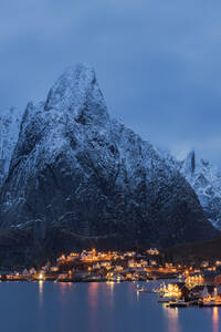 Spektakulärer Blick auf hohe schneebedeckte Berge und ein Dorf mit beleuchteten Häusern in einer Bucht am Fuße eines Hügels in Reine auf den Lofoten in Norwegen bei Sonnenuntergang - ADSF33706