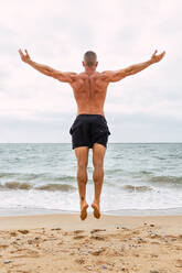 Ganzkörper-Rückenansicht eines unerkennbaren entschlossenen Sportlers mit nacktem Oberkörper, der hoch über der sandigen Küste in der Nähe des wogenden Meeres während des Trainings in der Natur springt - ADSF33715