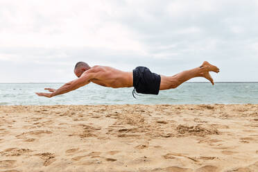 Ganzkörper-Seitenansicht eines gesichtslosen, hemdlosen Sportlers, der über die Sandküste springt, während er während des Trainings am Meer Liegestütze macht - ADSF33716