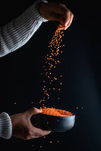 Hand of crop unrecognizable person pouring raw red lentils into bowl in black background - ADSF33718