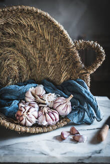 Cloves of unpeeled garlic served with blue napkin in round rattan basket placed on table in light kitchen - ADSF33721