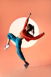 Seitenansicht einer energiegeladenen, fröhlichen Tänzerin, die mit erhobenem Bein und ausgestreckten Armen im Licht eines Projektors im Studio springt und wegschaut - ADSF33728