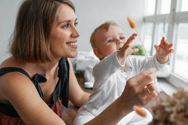 Fröhliche Mutter mit trockenen Pflanze spielen mit niedlichen kleinen Baby in weißer Kleidung in der Nähe von Fenster in hellen Raum zu Hause - ADSF33734