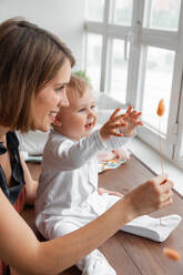 Fröhliche Mutter mit trockenen Pflanze spielen mit niedlichen kleinen Baby in weißer Kleidung in der Nähe von Fenster in hellen Raum zu Hause - ADSF33735