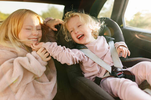 Positives Mädchen mit blonden Haaren spielt mit ihrer Schwester, die in einem schwarzen Kindersitz in einem modernen Auto sitzt, während einer gemeinsamen Autoreise - ADSF33738