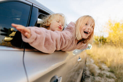 Niedliche Schwestern in Freizeitkleidung schauen aus dem Fenster, während sie im Auto sitzen, während einer gemeinsamen Autofahrt auf dem Land mit grünen Pflanzen - ADSF33740