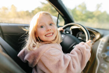 Seitenansicht eines fröhlichen Mädchens, das in die Kamera schaut und auf dem Fahrersitz eines modernen Autos sitzt und die Hände am Lenkrad hat - ADSF33741