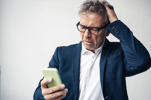 Frustrated businessman using smart phone against white background - JOSEF07300