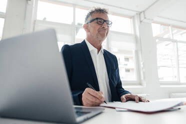 Smiling businessman with laptop and documents on desk in office - JOSEF07310