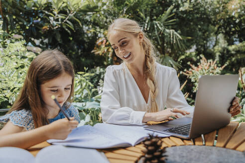 Lächelnde Mutter betrachtet ihre Tochter beim Lernen im Garten - MFF08587