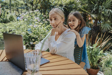 Smiling daughter watching mother using laptop in back yard - MFF08596