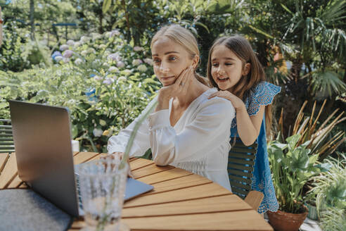 Lächelnde Tochter beobachtet Mutter bei der Benutzung eines Laptops im Hinterhof - MFF08596