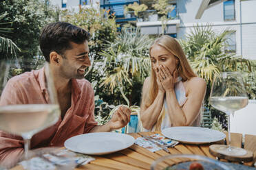 Surprised woman looking at man with engagement ring at outdoor table - MFF08617