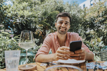 Happy man with mobile phone at outdoor table - MFF08621