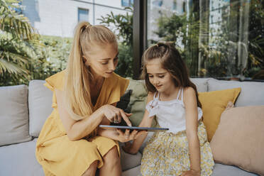 Mutter teilt Tablet-PC mit Tochter, die auf dem Sofa im Innenhof sitzt - MFF08632
