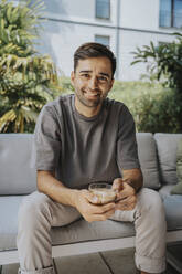 Smiling man holding coffee cup sitting in lounge - MFF08659