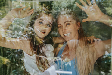 Smiling mother and daughter waving through window of house - MFF08665
