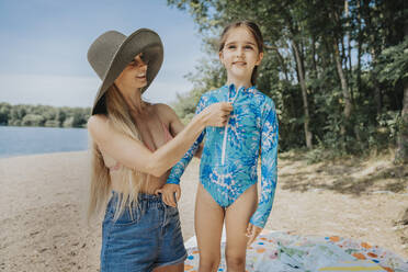 Mother helping daughter to wear swimwear at lakeshore - MFF08678