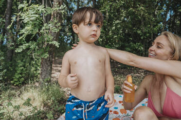Smiling mother applying sunscreen on son's back at lakeshore - MFF08728