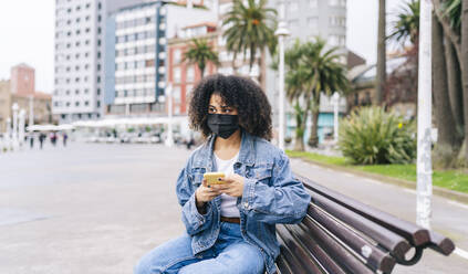 Woman wearing protective face mask with smart phone sitting on bench - DGOF02288