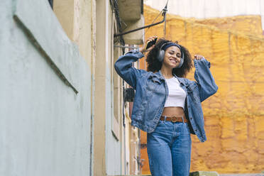 Happy woman listening music on headphones and dancing by wall - DGOF02311