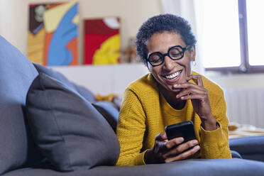 Smiling woman with eyeglasses using smart phone on sofa at home - EIF03444