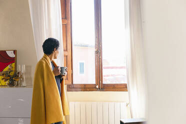 Woman wrapped in blanket holding mug and looking out of window at home - EIF03468