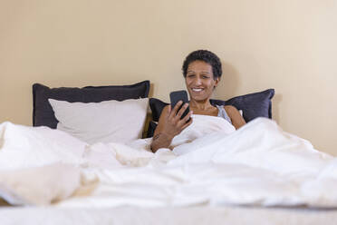 Smiling woman checking smart phone on bed at home - EIF03471