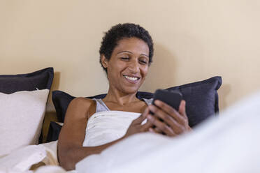 Smiling woman using mobile phone after waking up on bed at home - EIF03472