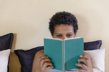 Woman with black short hair reading book on bed at home - EIF03475