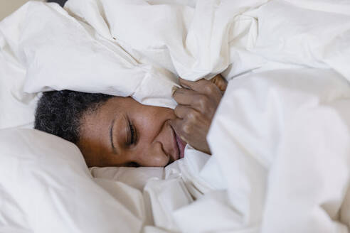Lächelnde Frau auf dem Bett liegend, bedeckt mit einer weißen Bettdecke, zu Hause - EIF03478