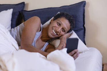 Smiling woman using mobile phone lying on bed in bedroom - EIF03481