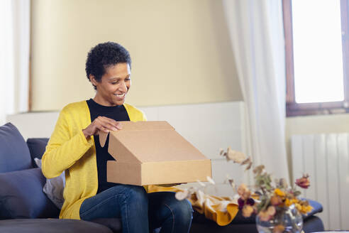 Woman opening cardboard box sitting on sofa at home - EIF03506