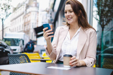 Lächelnde Geschäftsfrau mit Einweg-Kaffeebecher und Smartphone in einem Café - AMWF00190