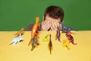 Man covering face with hands by dinosaur figurines against green background - IYNF00039