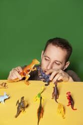 Junger Mann spielt mit Dinosaurier-Figur vor grünem Hintergrund - IYNF00042