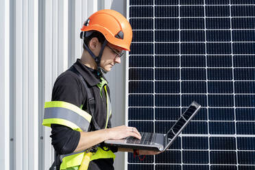 Junger Ingenieur bei der Arbeit an einem Laptop mit Sonnenkollektor in einem Solarkraftwerk - DLTSF02751