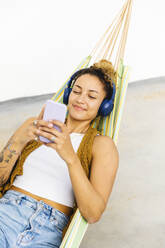 Smiling woman with headphones using smart phone lying on hammock - MRAF00849