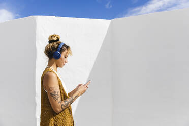 Woman with headphones using smart phone in front of wall on sunny day - MRAF00854