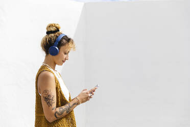 Young woman with headphones using smart phone in front of white wall - MRAF00855
