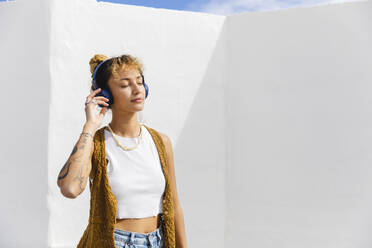 Hipster Frau mit geschlossenen Augen hört Musik über Kopfhörer vor einer Wand - MRAF00856