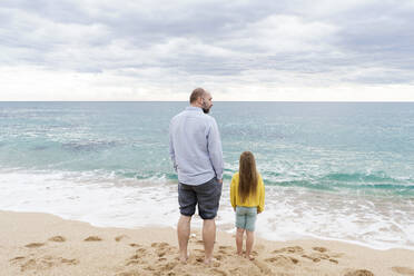 Wellen plätschern gegen Vater und Tochter, die am Strand stehen - KMKF01785