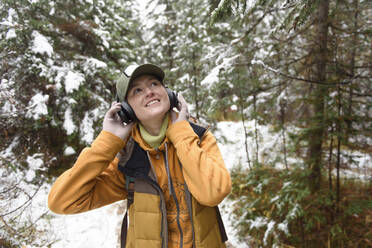 Smiling woman looking up listening music on headphones in winter forest - VBUF00014