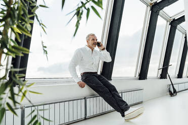 Businessman talking on mobile phone sitting on window sill at office - VPIF05441