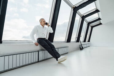 Businessman talking on smart phone sitting on window sill at work place - VPIF05442