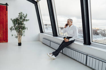 Geschäftsmann mit Einwegbecher, der in einem Startup-Büro mit seinem Smartphone telefoniert - VPIF05443