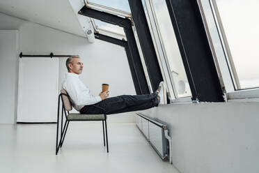 Geschäftsmann mit Einweg-Kaffeebecher sitzt auf Stuhl im Büro - VPIF05450