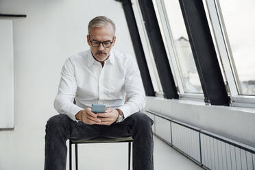 Geschäftsmann mit Brille, der im Büro eine Textnachricht auf seinem Mobiltelefon schreibt - VPIF05460